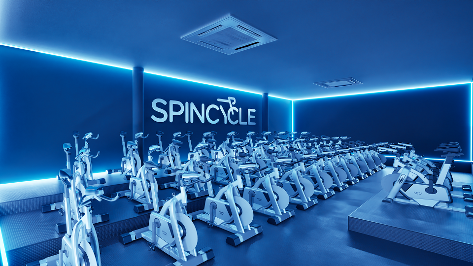 ala de ginástica com bicicletas ergométricas de spinning. A iluminação é azul e há fita ou luzes de LED ao redor do teto, criando um ambiente moderno. O nome “SPINCYCLE” está escrito na parede ao fundo, sugerindo que é um estúdio especializado em aulas de spinning.