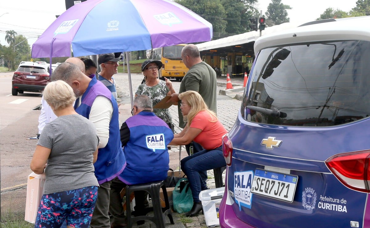 Carro do programa Fala Curitiba estacionado com guarda-sol do programa ao lado. Há pessoas vestidas com uniforme do programa conversando com outras de roupas comuns